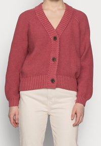 Prążkowany kardigan z dzianiny w głębokim różu z dekoltem w serek, czterema ciemnymi guzikami i balonowymi rękawami, zestawiony z jasnymi spodniami.