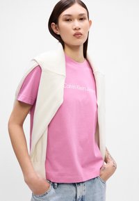 Gaišrozā kokvilnas T-krekls ar baltu "Calvin Klein Jeans" logotipu, apvienots ar krēma krāsas adītu džemperi, kas uzlikts pāri plecu zonai. Rādītas džinsu bikses.