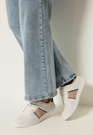 Baskets blanches avec des rayures beige et noires portées avec un jean large bleu clair, pieds croisés sur un fond clair uni.