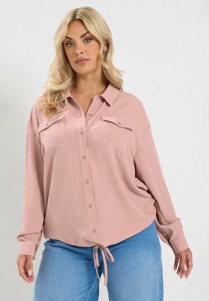 Blonde vrouw die een losse roze blouse met borstzakken en een blauwe spijkerbroek draagt, staand tegen een effen lichte achtergrond.