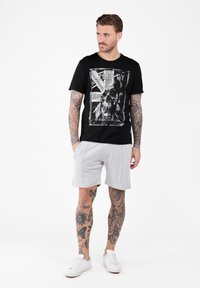 Mężczyzna z tatuażami na całej ręce i nodze, ubrany w czarny t-shirt z grafiką czaszki, jasnoszare spodenki i białe trampki, stojący z rękami w kieszeniach.