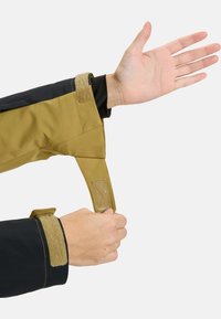 Der Ärmel der braun-schwarzen Jacke verfügt über einen verstellbaren Riemen mit Klettverschluss. Der Stoff ist glatt mit einem leichten Glanz und hat eine strukturierte Form.