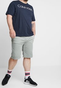 Laivaston sininen puuvillapaita, jossa on valkoinen "Calvin Klein" -logo, paritettu vaaleanvihreiden shortsien, mustien lenkkarien ja raidallisten valkoisten sukkien kanssa.