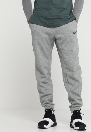 Homme portant un pantalon de survêtement gris Nike et des baskets Nike noir et blanc, debout les mains dans les poches devant un fond uni.