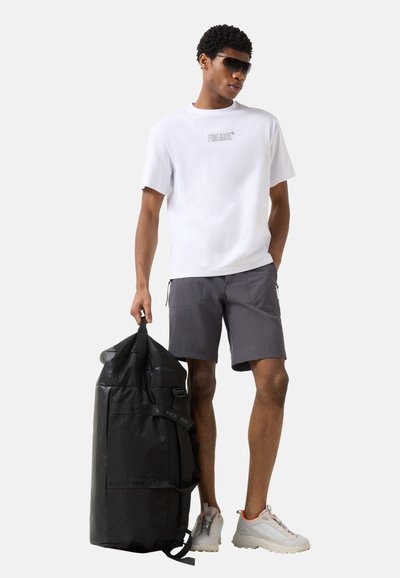 Homme portant des lunettes de soleil, chemise blanche à manches courtes, short gris, baskets blanches, tenant un grand sac de voyage noir à côté de lui.