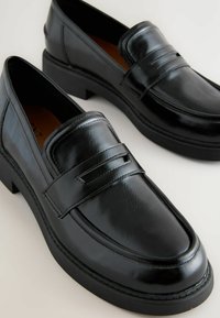 Mustat loaferit, jotka on valmistettu kiiltävästä nahasta, pyöreällä kärjellä, kohotetulla mustalla pohjalla ja koristeellisella nauhalla edessä.