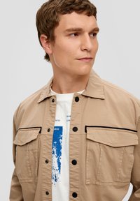 Giacca camicia beige, con bottoni e accenti neri, due tasche sul petto, presenta una texture liscia. T-shirt grafica bianca sotto.