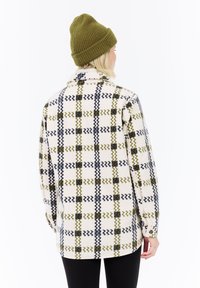 Persona con un gorro de punto verde y una chaqueta de forro polar blanca con un patrón de cuadros en zigzag verde y negro, de espaldas contra un fondo blanco.