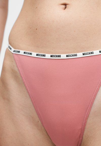 Gros plan sur la taille d’une personne portant un sous-vêtement rose avec une ceinture blanche comportant le texte noir répété « MOSCHINO ».