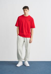 Rotes Baumwoll-T-Shirt mit schwarzem Logo, kombiniert mit grauen Jogginghosen und weißen Sneakern. Schlichter Hintergrund mit blauem Boden. Lässiges Outfit.