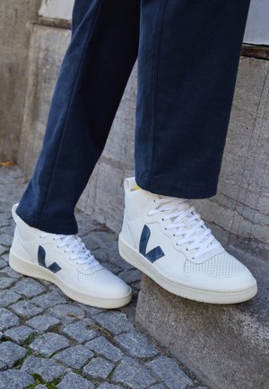 Zapatillas altas blancas con logo V azul marino usadas con pantalones azul oscuro en calle empedrada y escalón de piedra.