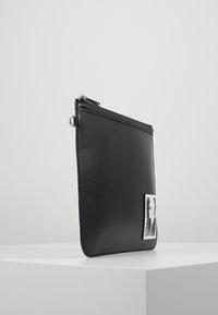 Černé kožené psaníčko s hladkou texturou, zapínáním na zip a stříbrným kroužkem jako ozdobou. Na přední straně se nachází malý grafický potisk.