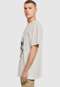 Lys grå t-shirt med korte ærmer, der har en stor sort kraniegrafik på fronten. Fremstillet af bomuld med en afslappet pasform.