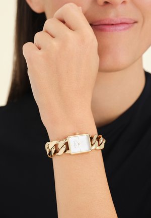 Montre en or avec un cadran rectangulaire blanc, bracelet en chaîne à maillons, et un design minimaliste. Portée sur un poignet de teinte de peau claire.