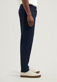 Marineblauwe, taps toelopende broek met een elastische tailleband, zijzakken en een gladde textuur. Gepaard met witte sneakers met bruine zolen.