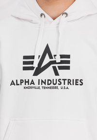 Sudadera blanca con cordones grises, que presenta un logo impreso en negro y el texto "ALPHA INDUSTRIES" y "KNOXVILLE, TENNESSEE, U.S.A."