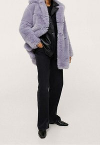 Persona che indossa un cappotto di pelliccia sintetica lavanda sopra una camicia di pelle nera, jeans neri e stivali a punta nera su uno sfondo neutro.