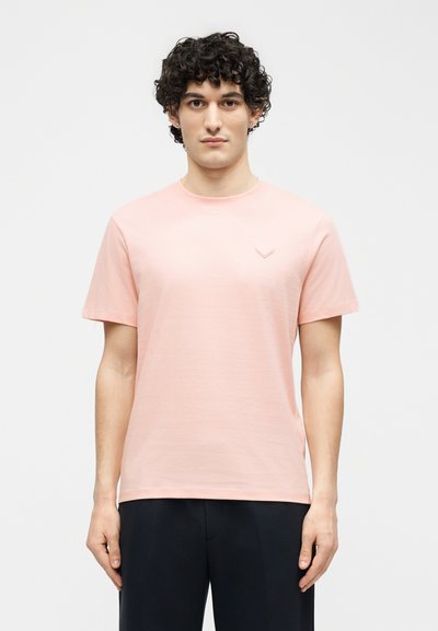 Νέος άνδρας με σγουρά μαλλιά, φορώντας ένα απλό ροζ κοντομάνικο μπλουζάκι και μαύρο παντελόνι, στέκεται μπροστά σε λευκό φόντο.