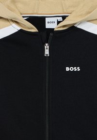 Μαύρο φούτερ με φερμουάρ, με μπεζ λεπτομέρειες και λευκές ρίγες. Διαθέτει ασημένιο φερμουάρ και λευκό λογότυπο "BOSS" στην περιοχή του στήθους.