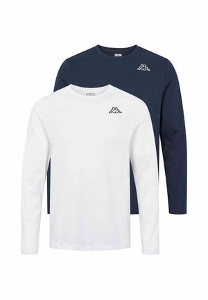 Muž nosící tmavě modrou košili s dlouhým rukávem s malým bílým logem, volné světle modré džíny a bílé tenisky, stojící před jednobarevným pozadím.