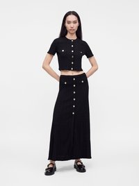 Femme portant un haut court noir à manches courtes avec boutons, et une jupe longue boutonnée assortie, avec des chaussures noires sur fond blanc.