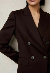 Blazer double boutonné marron foncé avec revers crantés, deux boutons, une coupe ajustée et poches latérales, confectionné en tissu lisse.