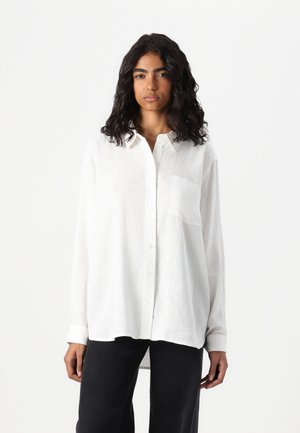 White linen button-up shirt with long sleeves, a single chest pocket, and a relaxed fit that extends slightly at the back.