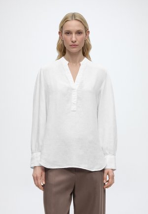 Žena s dlhými blond vlasmi, oblečená v bielu blúzku s dlhým rukávom a výstrihom do V a hnedé nohavice, stojaca pred jednoduchým pozadím.