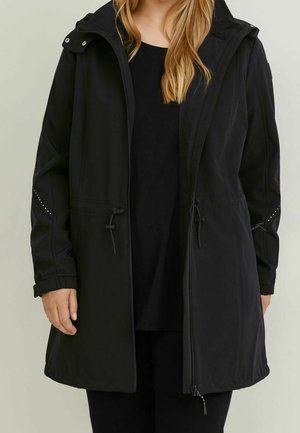 Une femme porte un long manteau noir à capuche, avec une fermeture éclair sur le devant, une taille à cordon de serrage et des détails cloutés aux manches, par-dessus des vêtements noirs.