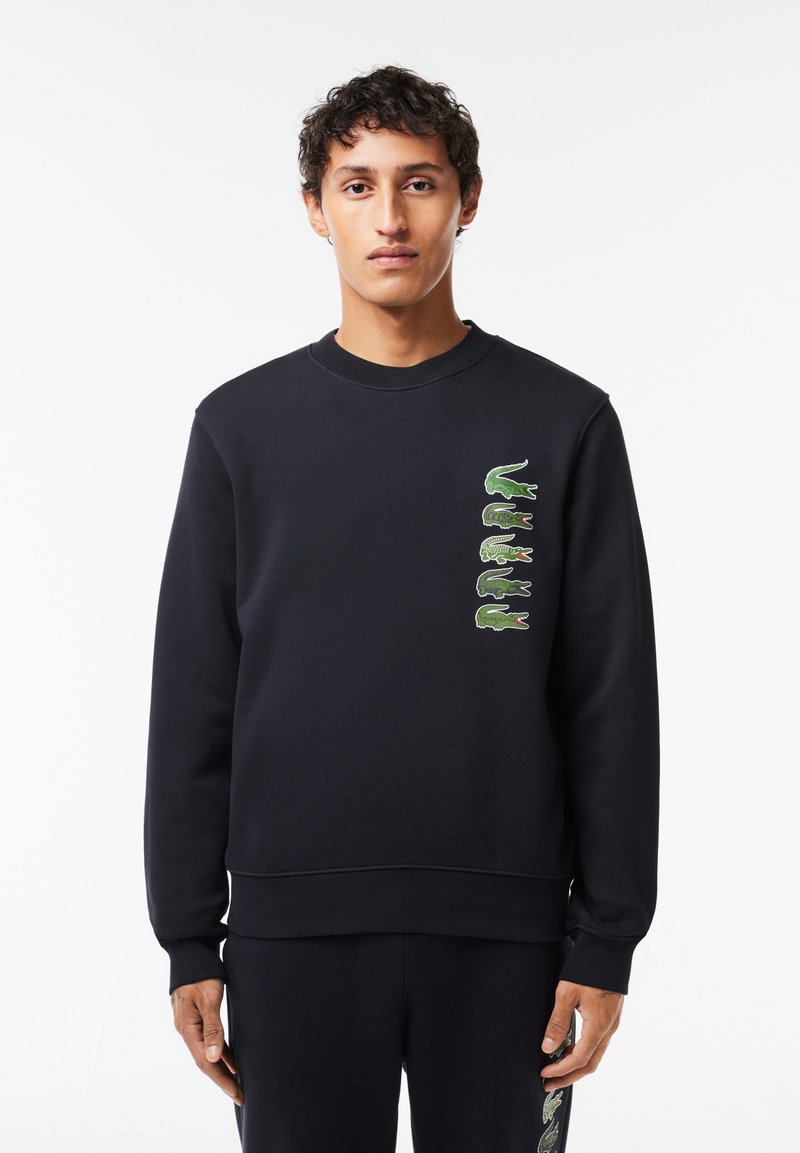 Sudadera negra de algodón con puños y dobladillo acanalados. Presenta logotipos de cocodrilo verdes en varias poses a lo largo del lado izquierdo.