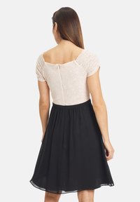 Kobieta ubrana w sukienkę w dwóch kolorach, z jasnoróżową koronkową górą i krótkimi rękawami oraz czarną spódnicą do kolan, odwrócona plecami do kamery.