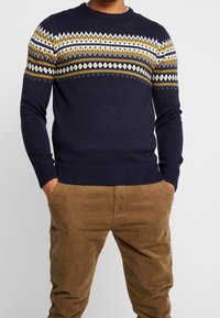 Navy knitted sweater with a geometric yellow, white, and grey design across the chest, paired with tan corduroy pants.
