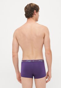 Mann steht mit dem Rücken zur Kamera, trägt lila Boxershorts mit einem Taillengummi mit "sloggi"-Logo vor weißem Hintergrund.