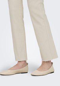 Piedi che indossano ballerine beige a punta abbinate a pantaloni dritti color crema con orli sfrangiati.
