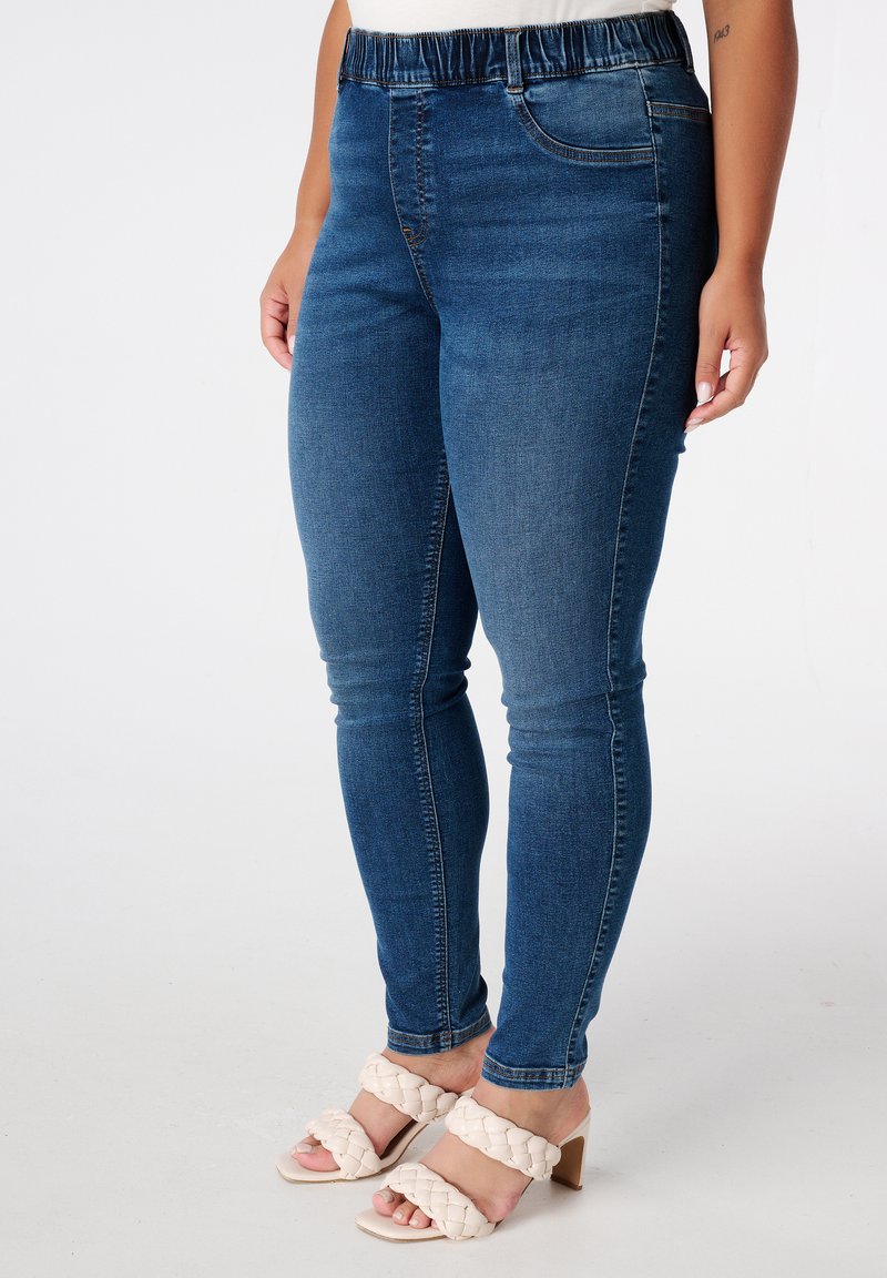Jeggings en denim bleu avec une taille élastique et deux poches avant. Associées à des sandales à talons hauts à lanières tressées crème. Texture lisse.