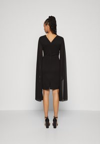 Μαύρο φόρεμα με μακριά, διαφανή μανίκια, σχέδιο V στην πλάτη, μήκος μέχρι το γόνατο και κεντρικό φερμουάρ στην πλάτη, φορεμένο με μαύρες ψηλές γόβες.