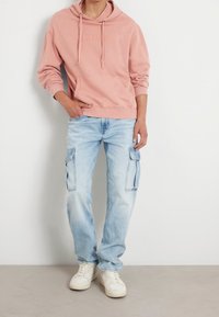 Sudadera con capucha de color rosa claro con bolsillo frontal y logo bordado, combinada con pantalones cargo de denim azul claro que tienen bolsillos laterales y zapatillas blancas.