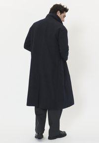 Garšs tumši zilā krāsā mētelis ar augstu apkakli, aizmugures krokām un strukturētām pleciem; apvienots ar tumšām biksēm un melniem stingriem kurpēm.