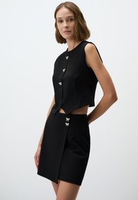 Schwarzes ärmelloses Crop-Top und Minirock-Set aus glattem Stoff. Ausgestattet mit silbernen dekorativen Knöpfen und einem Ausschnitt an der Seite.