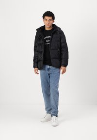 Chaqueta acolchada negra con capucha y cierre de cremallera, con un diseño acolchado. Combinada con una camisa negra, jeans de mezclilla azul claro y zapatillas blancas.