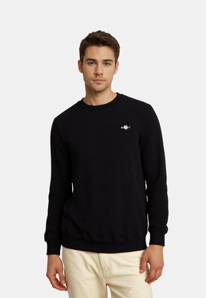 Czarny teksturowany sweter z okrągłym dekoltem, z prążkowanymi mankietami i dołem, z małym haftowanym logo na piersi. Noszony z jasnymi spodniami.