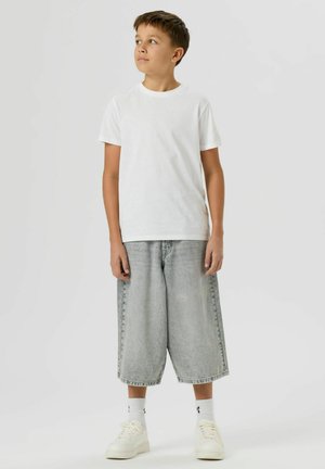 Chlapec stojí a dívá se vzhůru, má na sobě bílé tričko, dlouhé šedé džínové šortky, bílé tenisky a bílé kotníkové ponožky na jednobarevném pozadí.