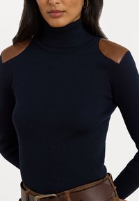 Pull à col roulé côtelé navy avec des accents en cuir brun sur les épaules, présentant une coupe ajustée et une texture détaillée.