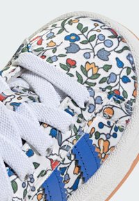 White sneaker featuring a floral pattern in red, blue, and green, with white laces, blue leather accents, and a textured rubber sole.