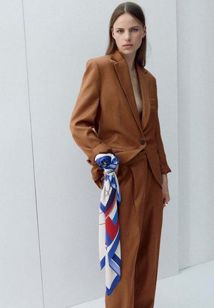 Femeie purtând un costum maro cu un nasture, ținând o eșarfă cu model albastru, alb și roșu pe un fundal simplu gri deschis.