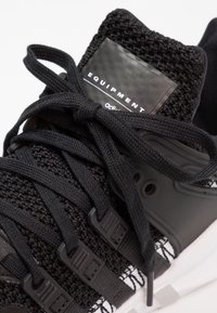 Svart idrottssko med texturerad stickad ovandel, tjocka snören, plastdetaljer och en framträdande logotypens patch på tungan.