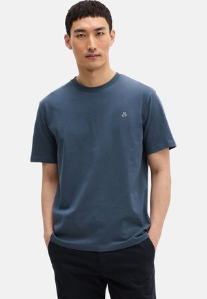 Man draagt een donkerblauw T-shirt met korte mouwen en kleine witte tekst op de borst, staat met handen in de zakken tegen een egale achtergrond.
