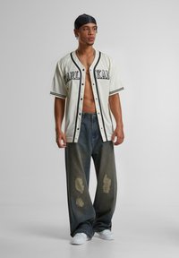 Cămașă albă cu butoni, având accente negre și logo, asortată cu pantaloni evazați din denim închis, cu un aspect uzat. Modelul poartă adidași albi.