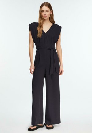 Frau trägt einen ärmellosen schwarzen Jumpsuit mit V-Ausschnitt, weitem Bein und passendem Gürtel, kombiniert mit schwarzen Plateausandalen.