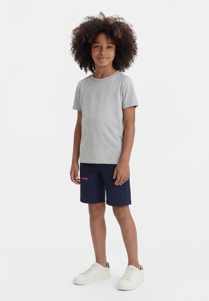 Copil cu păr creț, zâmbind, purtând un tricou gri cu mâneci scurte, pantaloni scurți bleumarin cu buzunar cu fermoar și adidași albi, pe un fundal alb.
